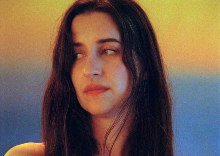 Portret van een vrouw met lang, donker haar, die in de camera kijkt tegen een kleurrijke achtergrond.