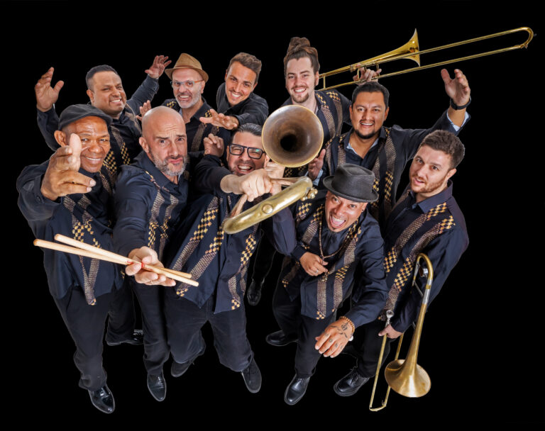 Een groep van negen muzikanten poseert samen met verschillende blaasinstrumenten op een zwarte achtergrond.