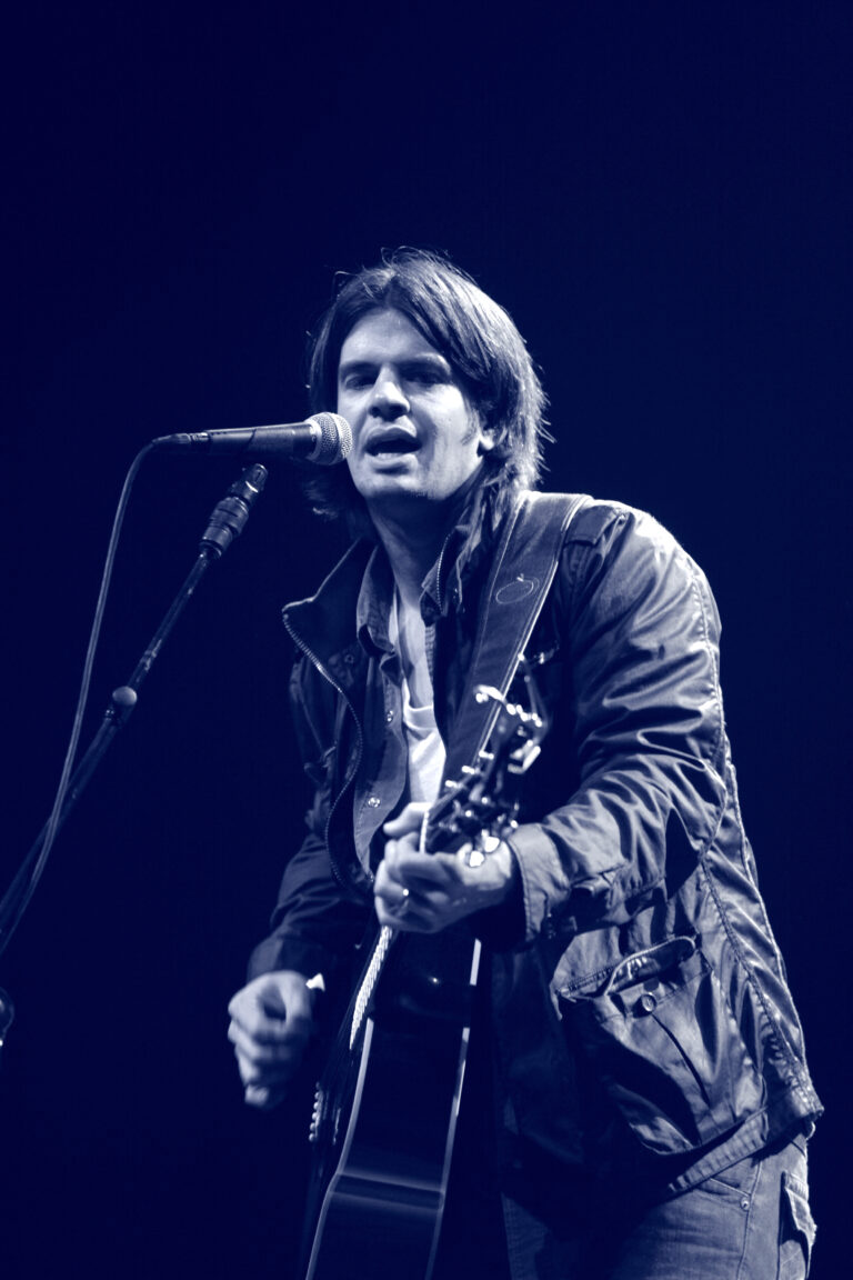 Een muzikant die een gitaar speelt en in een microfoon zangt, met een blauwe achtergrond.