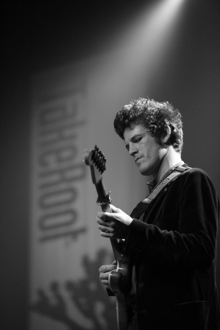 Een muzikant speelt gitaar op een podium met een logo op de achtergrond.
