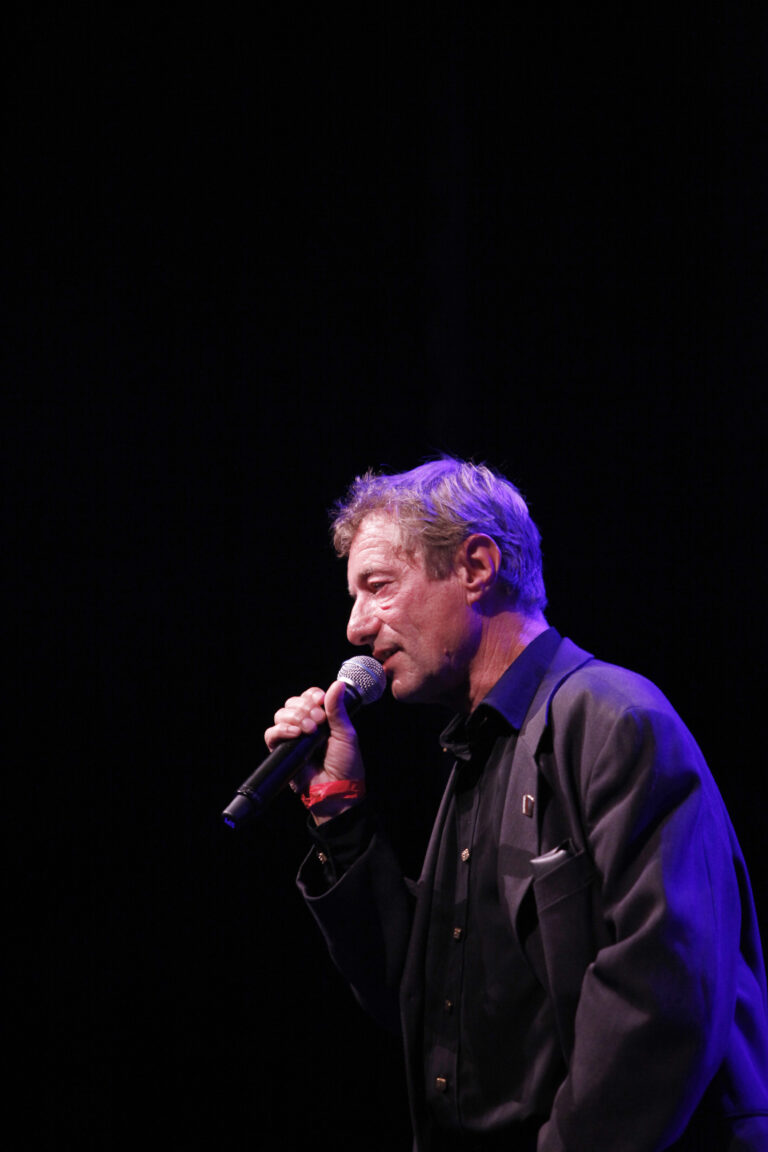 Een man spreekt in een microfoon op een podium met een donkere achtergrond en paarse verlichting.