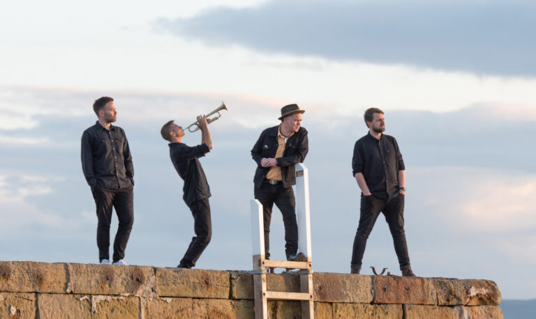 vier mannen in zwarte kleding naast elkaar op een muurtje waarvan een met een trompet