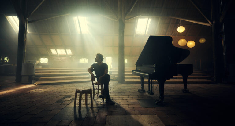 Man zit op stoel in grote donkere schuur met piano en binnenvallend licht.