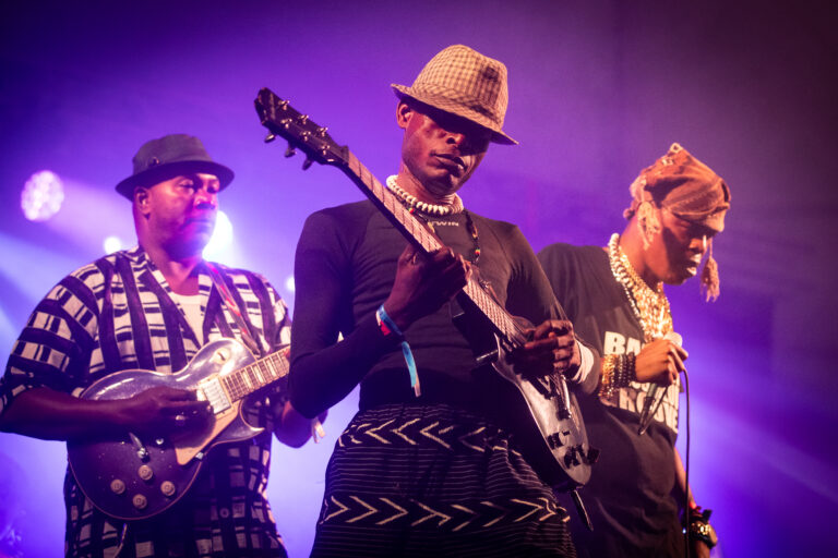 Drie mali muzikanten met twee gitaren en paarse achtergrond.