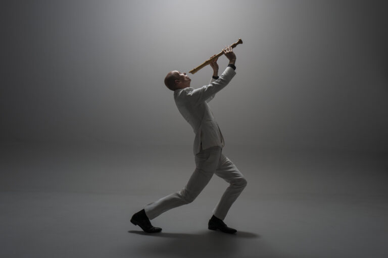 Blokfluitist van zijn rechterzijkant gefotografeerd, gekleed in wit pak en zwarte schoenen in licht achterovergebogen houding zijn hoofd omhoog tegen een grijze achtergrond. In zijn handen een middelgrote blokfluit die net niet zijn mond raakt.