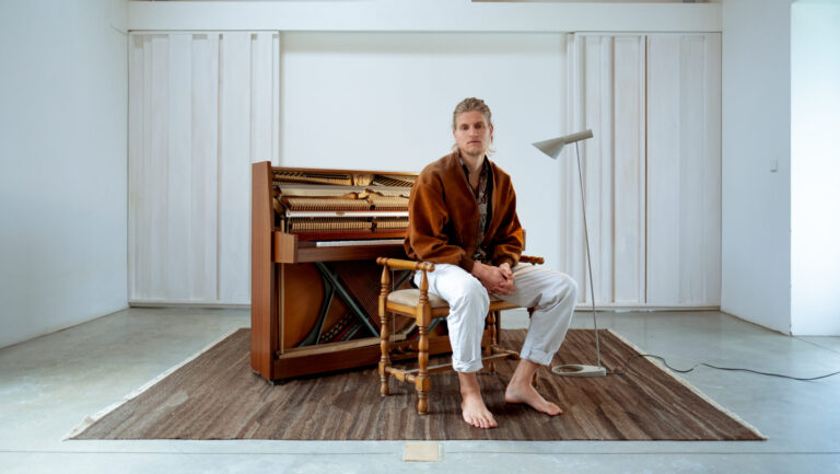 Challem zit met blote voeten voor een houten piano en de achtergrond is een witte muur