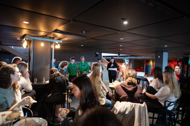 Studenten zitten in het cafe aan de tafels en er wordt verteld door een presentator