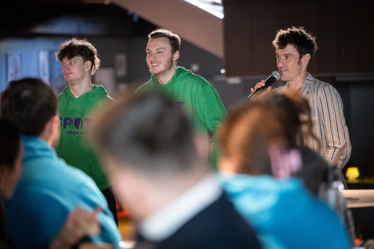 Drie personen waarvan twee met felgekleurde groene trui aan. De derde persoon heeft een microfoon