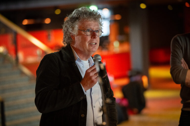 Man met bril en zwart jasje en overhemd presenteert met microfoon