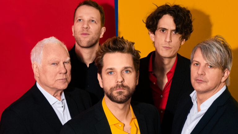 Foto van 5 mannen in zwarte pakken voor een geel met rode muur, de band Sloper.