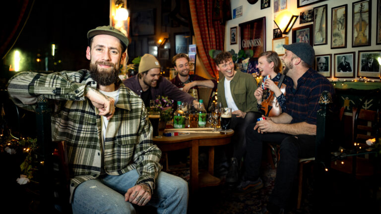 TikTok Tammo zit op de voorgrond in de kroeg, achter hem zit een groepje mensen breed lachend wat te drinken