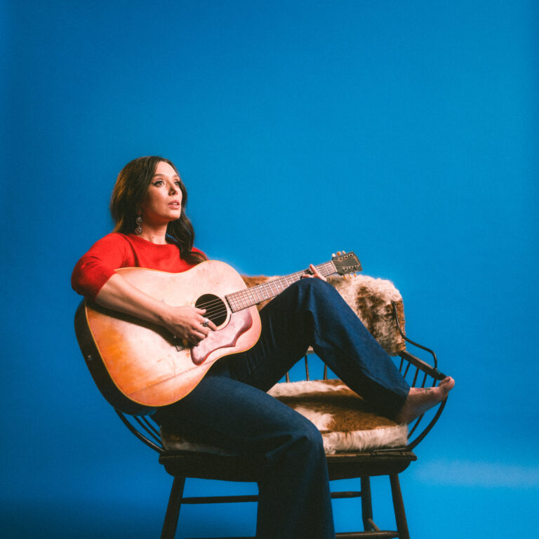 Vrouw met rode trui zit op een stoel met een gitaar voor een blauwe achtergrond