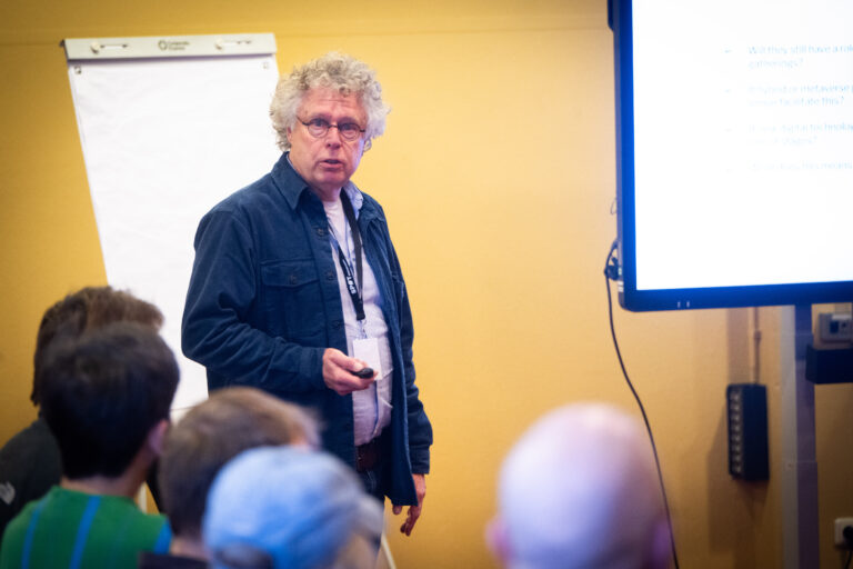 Een man met donkerblauw vest voor een gele muur