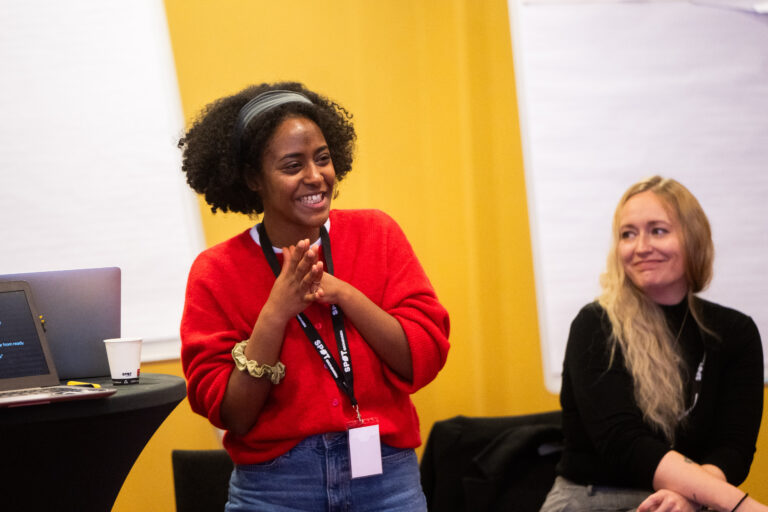 Een vrouw in een rode trui met haar handen in elkaar voor een gele muur