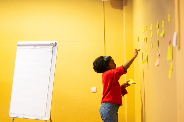 Een vrouw met oranje trui schrijft op post it en staat voor een gele muur