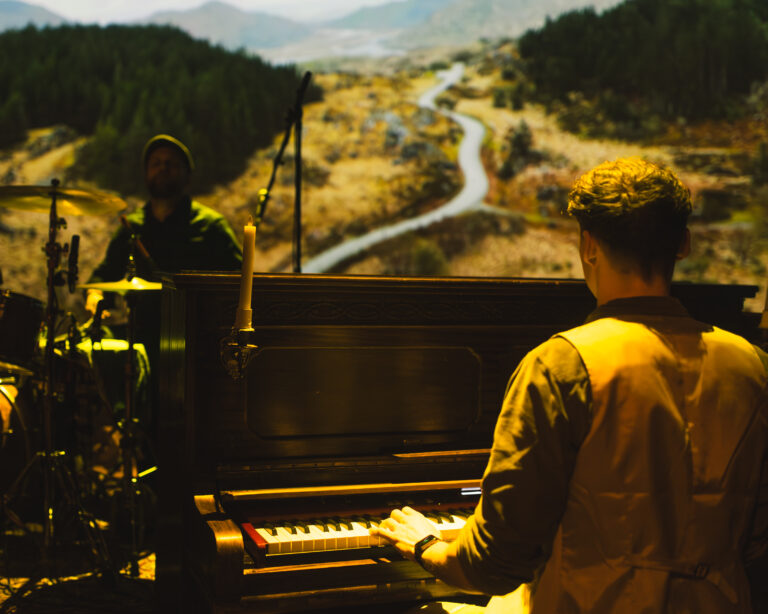Twee muzikanten spelen een piano op een podium, met een lange landweg in de achtergrond.
