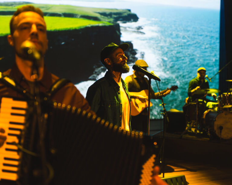 Een groep muzikanten speelt buiten aan de kust, met de oceaan op de achtergrond.