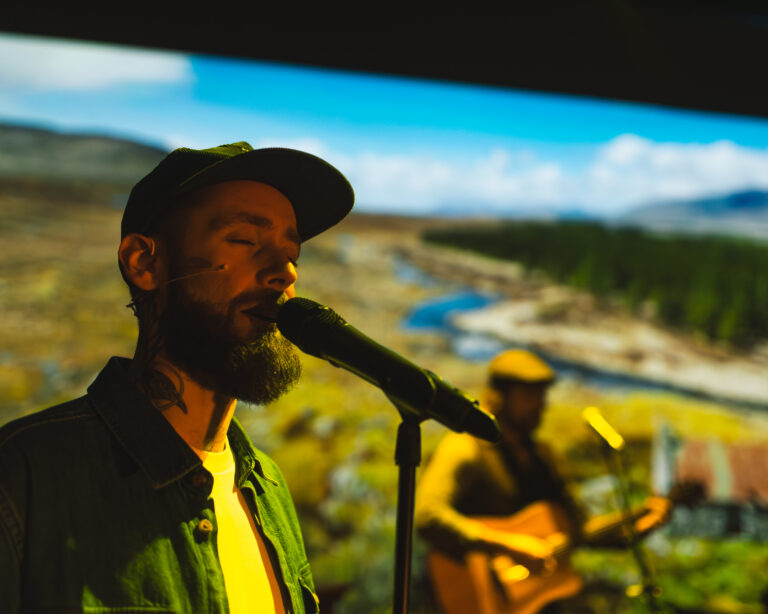 Een man met hoed zingt in een microfoon tijdens een buitenoptreden, met een gitarist op de achtergrond.