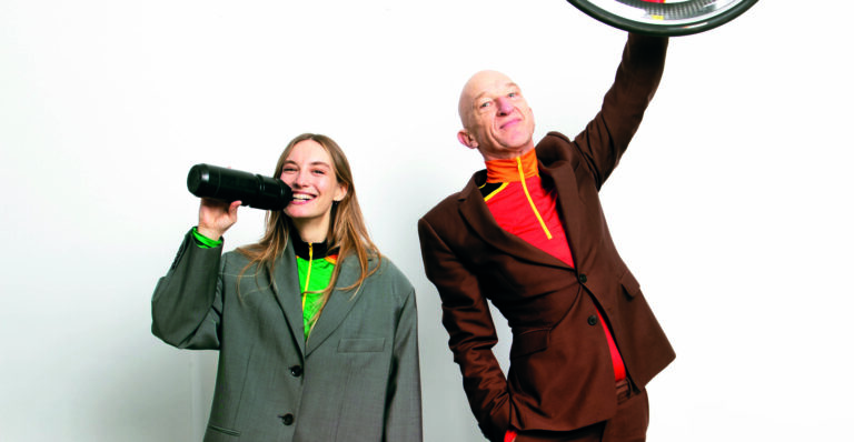Caro Derkx en Wilfried de Jong staan allebei in wielrentenue met een net pak eroverheen. Caro met haar mond aan een bidon en Wilfried steekt een fietswiel omhoog.