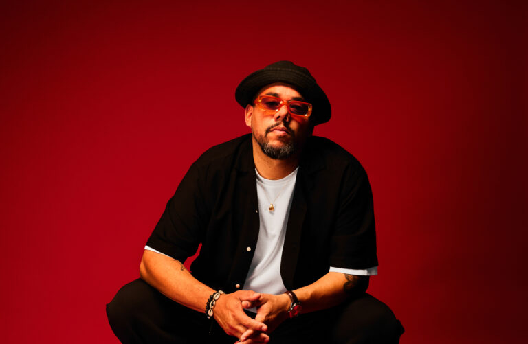 Een man met een zwarte pet en een open zwarte jas over een witte T-shirt zit tegen een felrode achtergrond.