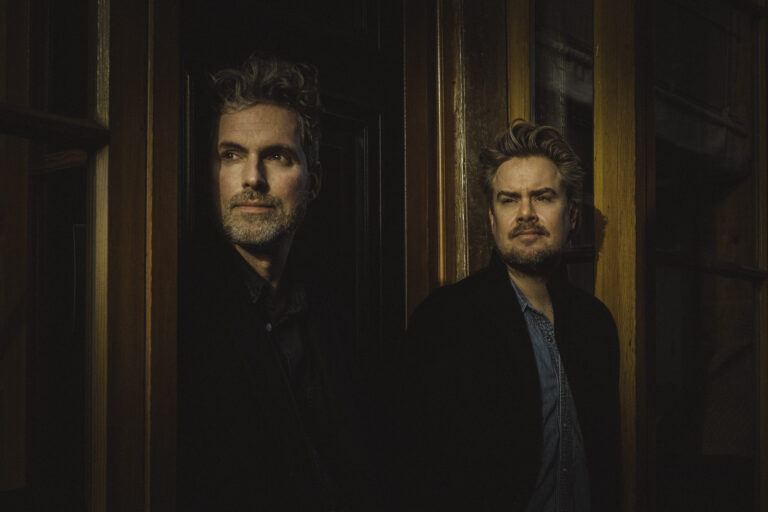 Twee mannen staan in een donkere ruimte met houten elementen, waarbij één van hen lichtjes naar de camera kijkt.