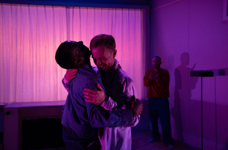 Twee mensen omhelzen elkaar voor paars licht, met een persoon op de achtergrond.