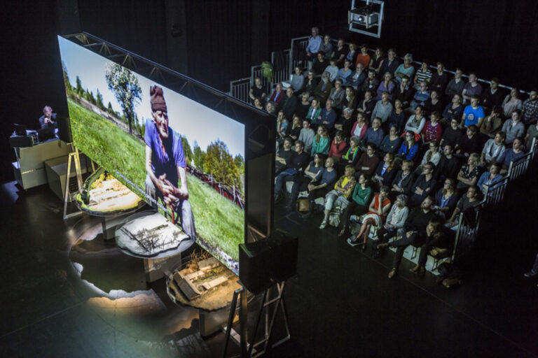 Publiek kijkt naar groot scherm met daarop een projectie van een oude man
