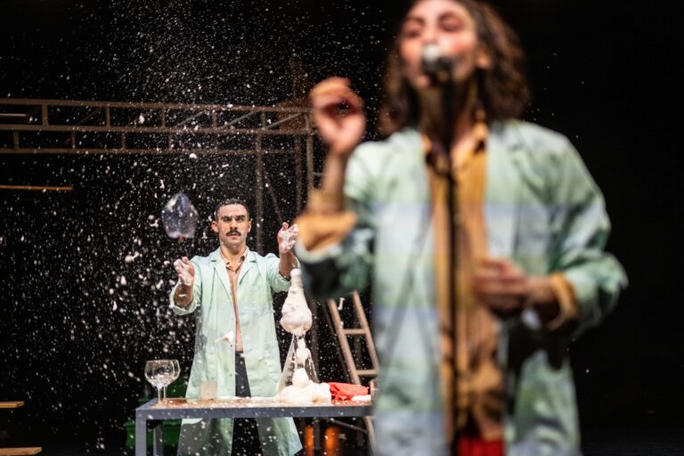 Twee artiesten op een podium, met één die iets uit een glas laat vallen en een andere die spreekt in een microfoon, terwijl er een sneeuwachtige substantie rondom hen valt.