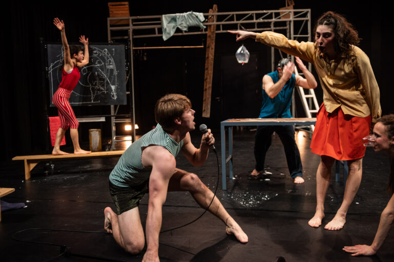 Een toneelopvoering met vijf acteurs in verschillende posities, waarbij één acteur op de grond met een microfoon zingt, terwijl anderen actief deelnemen aan de scène.