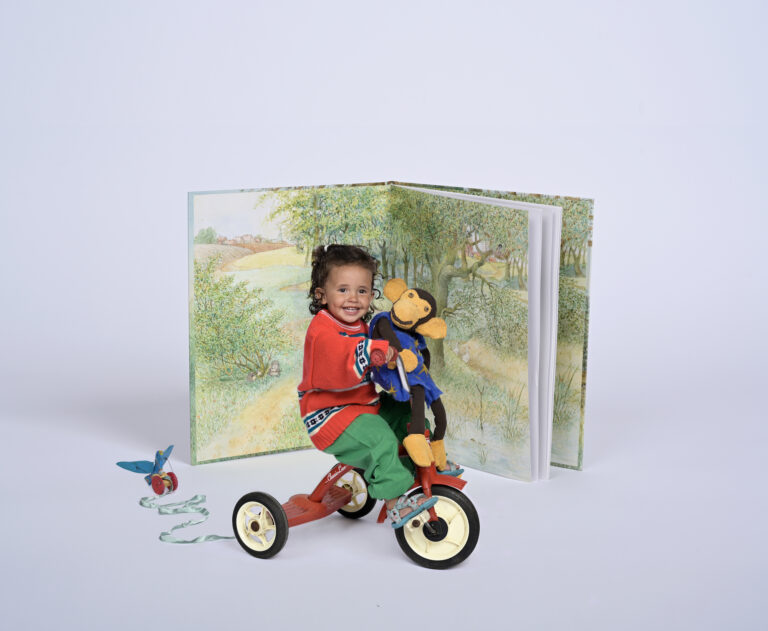 Een kind zit op een driewieler en houdt een speeltje vast, met een grote open kinderboek op de achtergrond.