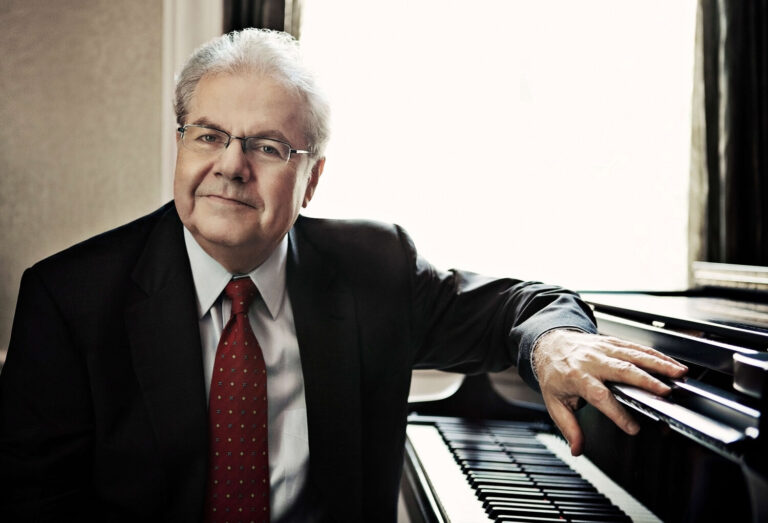 Emanuel Ax in pak met rode stropdas achter een piano kijkt de camera in