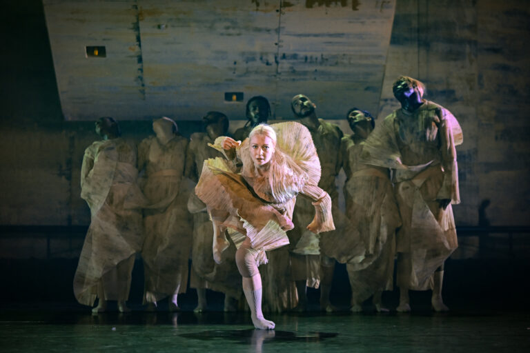 danseres met opwaaiende jurk staat op één been en kijkt recht in de camera