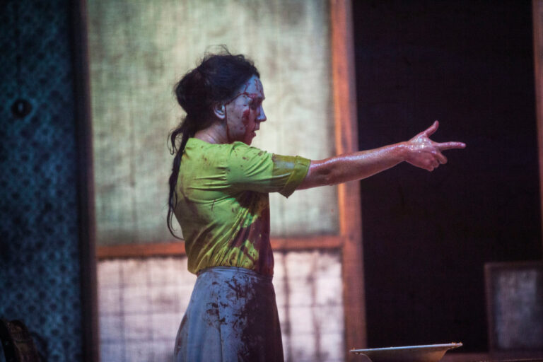 Een bebloede vrouw maakt een schietbeweging met haar arm