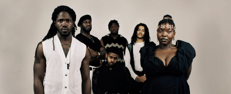 5 mensen zwart wit gekleed voor witte achtergrond staan stoer geposeerd om man op stoel.