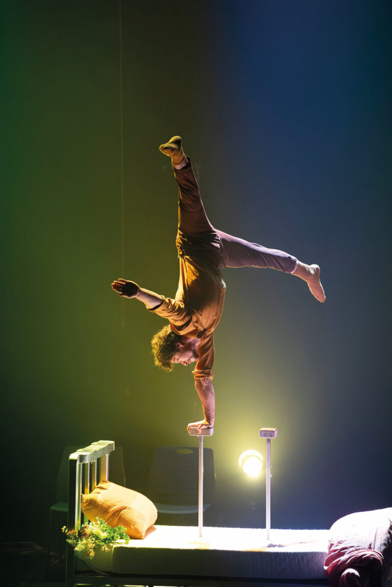 Een acrobaar balanceert op 1 hand op een bed met donkere achtergrond.