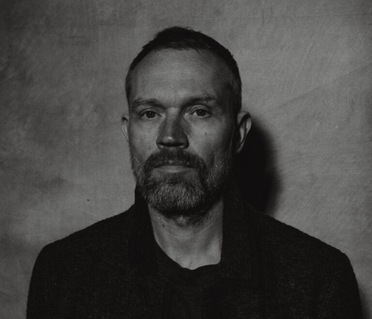 Zwart/wit foto van man met kort haar en ringbaard in donkere kleding.