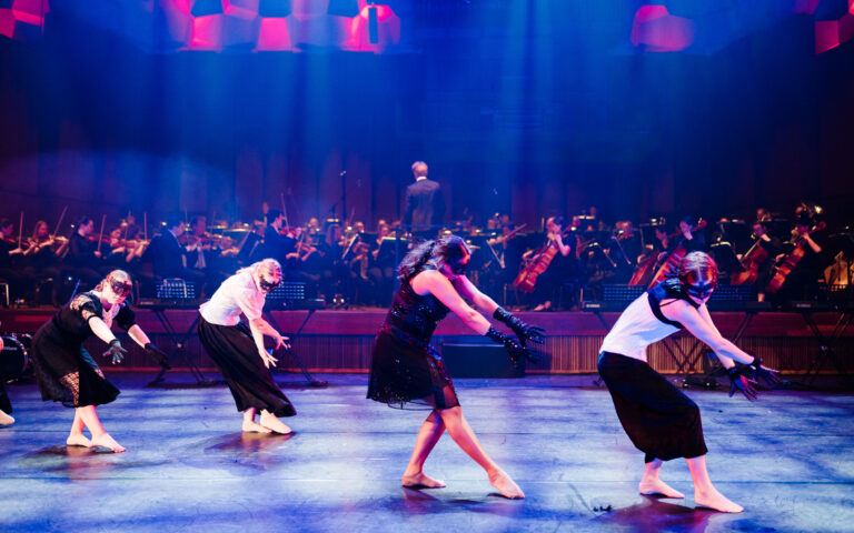 Op de voorgrond vier gemaskerde danseressen, op de achtergrond het Noord Nederlands Orkest