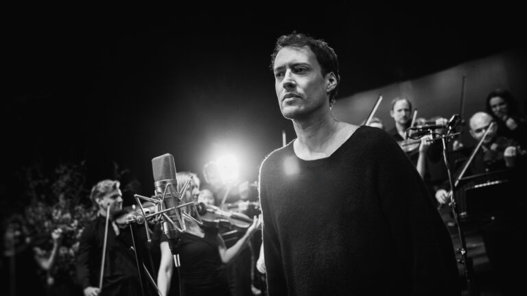 Zwart-witte foto van man met microfoon, achter hem staat een orkest