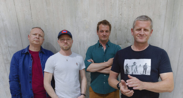 Vier leden van Racoon poseren staand voor een witte houten wand