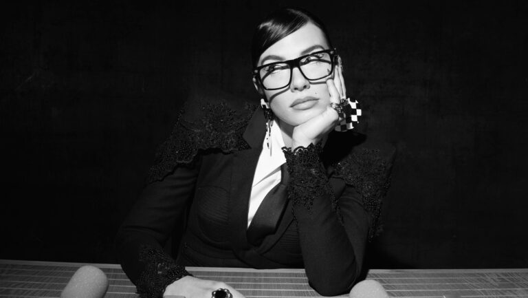 Vrouw met grote zwarte bril zit achter bureau met microfoons