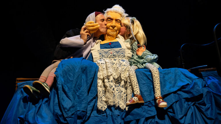 Drie mensen in historische kostuums op een podium, met een centraal figuur in een glanzende jurk en witte pruik.