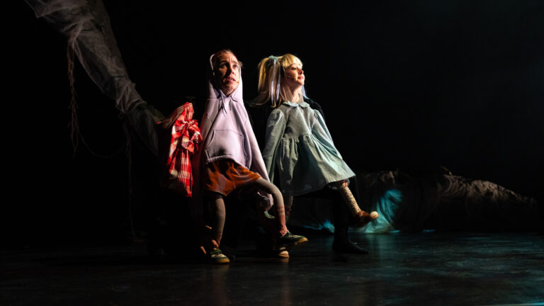 Drie dansers in kleurrijke outfits bewegen op een donkere scène onder een spotlicht.