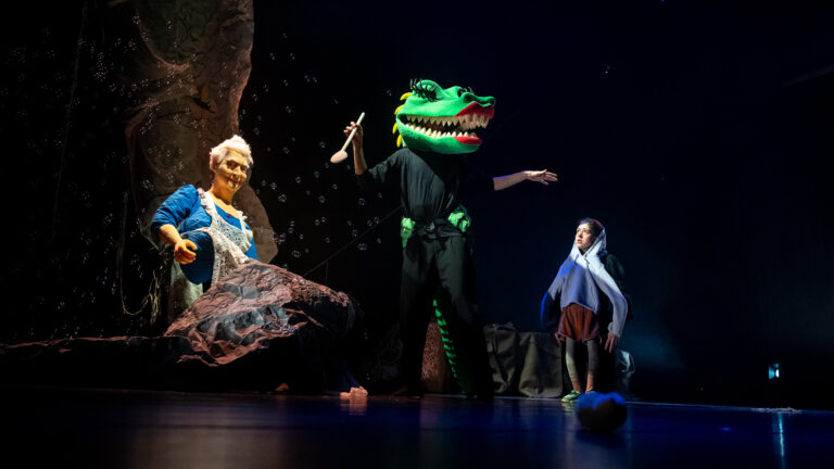 Een groen draakachtig figuur staat centraal op een donker podium, met twee acteurs in kostuums aan weerszijden.