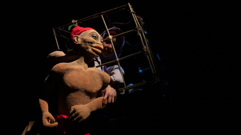 Een performer in een bruine vachtkostuum met een rode hoed staat in een metalen kooistructuur tegen een donkere achtergrond.