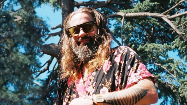 Man met een lange baard en zonnebril, gekleed in een kleurrijke blouse, staat buiten bij boom.