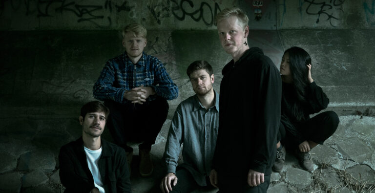 Donkere mysterieuze foto van 1 man staand, 3 mannen zittend en 1 vrouw zittend op stenen onder een brug.