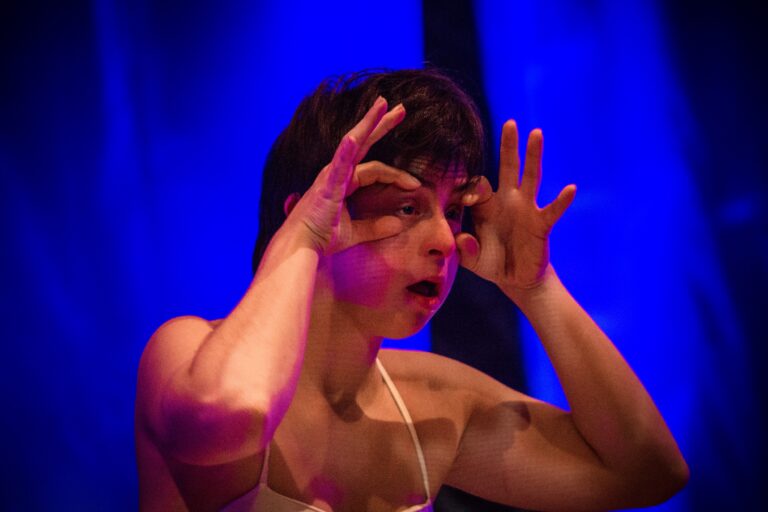 Een performer in een wit kostuum maakt een expressieve gezichtsuitdrukking met de handen voor de ogen, tegen een blauwe achtergrond.