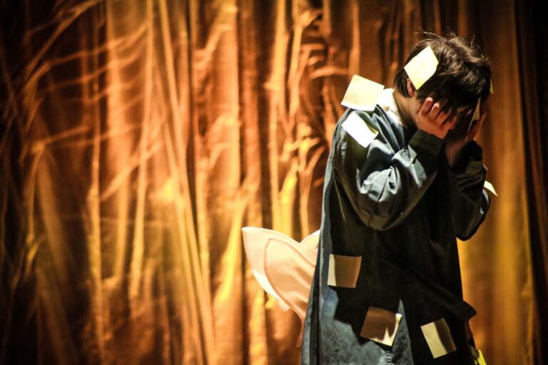 Een persoon in een spijkerjack met papieren briefjes op hun kleding staat met de handen voor hun gezicht op een podium met een oranje achtergrond.