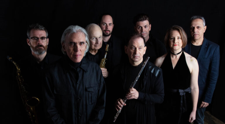 8 muzikanten, waarvan 1 vrouw, staan in zwarte kleding voor een zwarte achtergrond met instrumenten in handen.