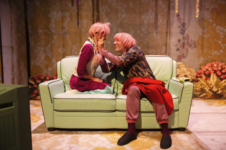 Twee personages op een groene bank hebben allebei een roze pruik op, de een knijpt de ander in de wangen